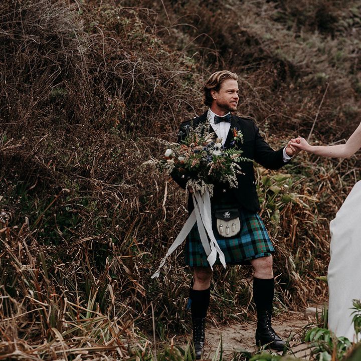 Groom in a blue and green tartan kilt holding his brides hand in a April Ross wedding dress and her boho wedding bouquet 