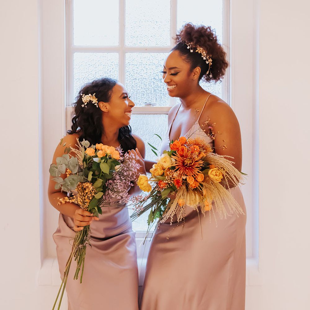 Bridesmaids in dusky rose satin bridesmaid dresses holding yellow and orange flower bouquets and wearing jewelled hair accessories 