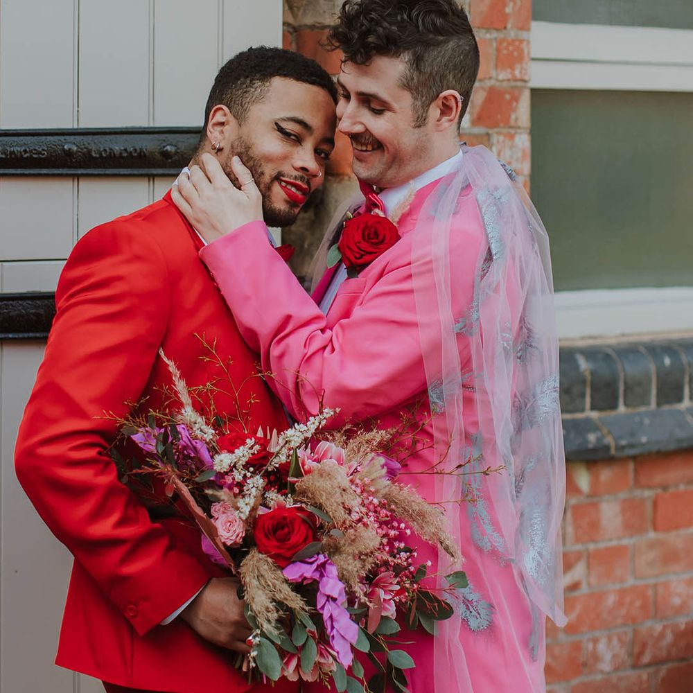 Stylish groom in a pink two piece suit with bow tie, and embroidered wedding veil embracing his partner in a red suit holding a pink wedding bouquet 