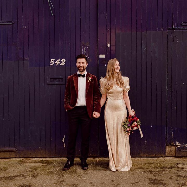 Bride in gold puff sleeve wedding dress with bow detail intertwines her fingers with the groom in red suit jacket 