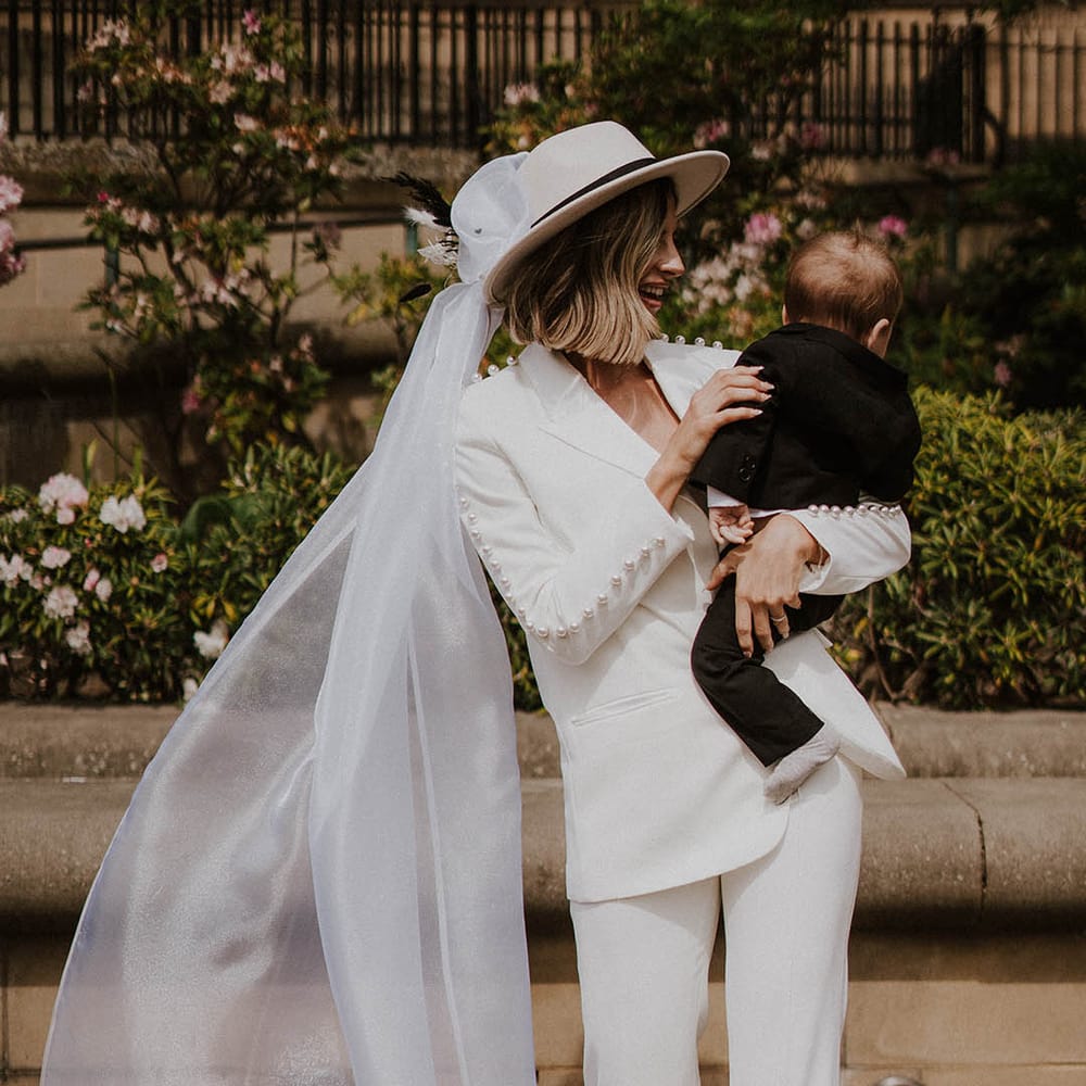 Bride with bobbed hair wearing white Nadine Merabi pearl trouser suit with veil attached holding son in black tie 