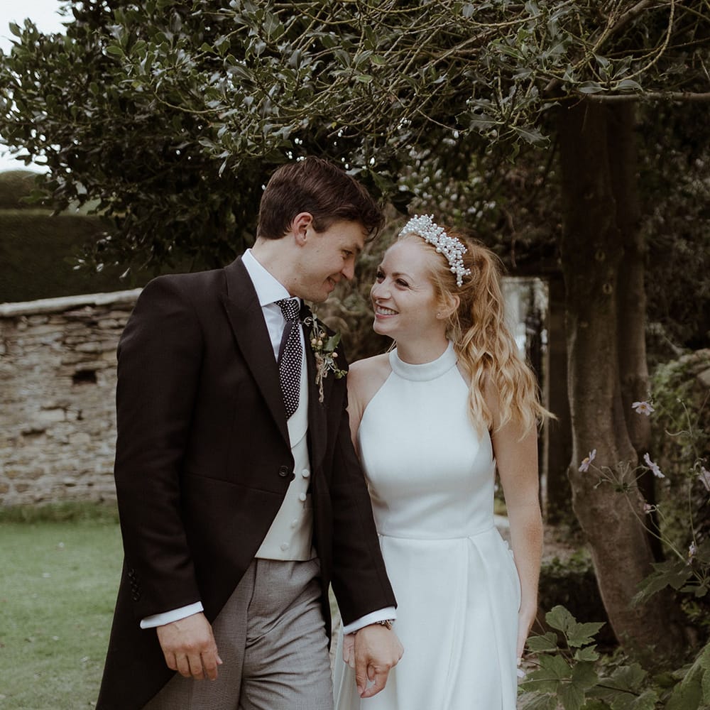 Bride and groom hold hands as they star at each other for their couple portraits with groom in morning suit and grey trousers and bride in a halter neck wedding dress 