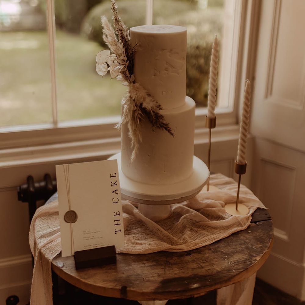 Two tier white wedding cake with dried grass decoration, twisted candles and a cake sign with a wax seal