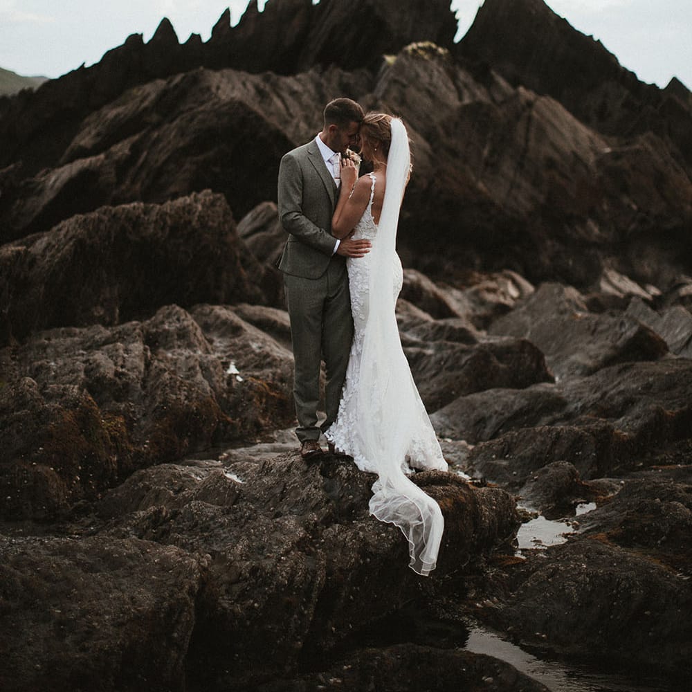 Bride wears low back fitted Enzoani wedding dress and floor length veil as she embraces her groom on Tunnels Beach surrounded by rocks 