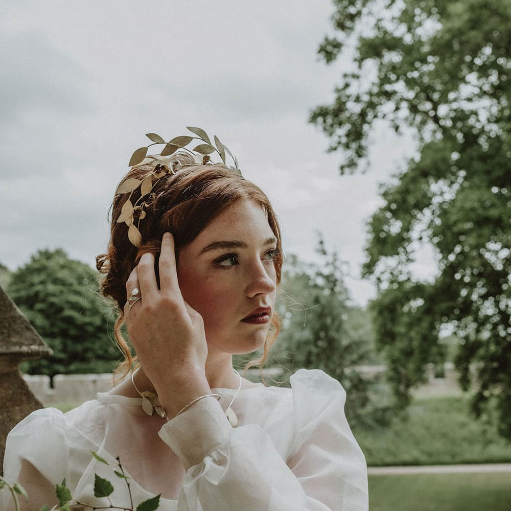 Bride wearing a sheer long sleeve wedding dress and gold leaf headpiece holding an oversized bridal bouquet with green foliage and white and yellow flowers 
