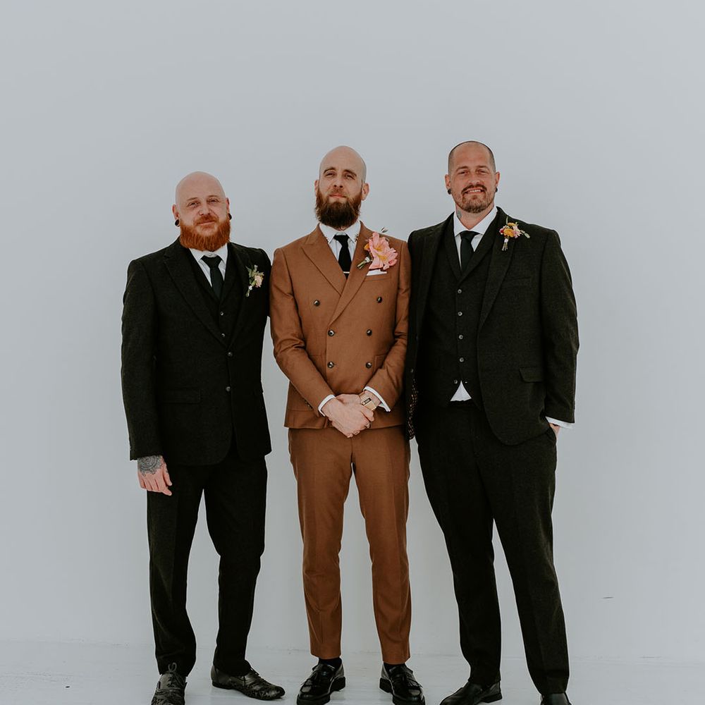 Groom wears brown retro styled suit with statement floral buttonhole and stands with his groomsmen in dark suits 