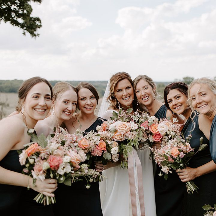 Bride poses with her bridesmaids wearing one shoulder green satin bridesmaid dresses and holding pastel bouquets 