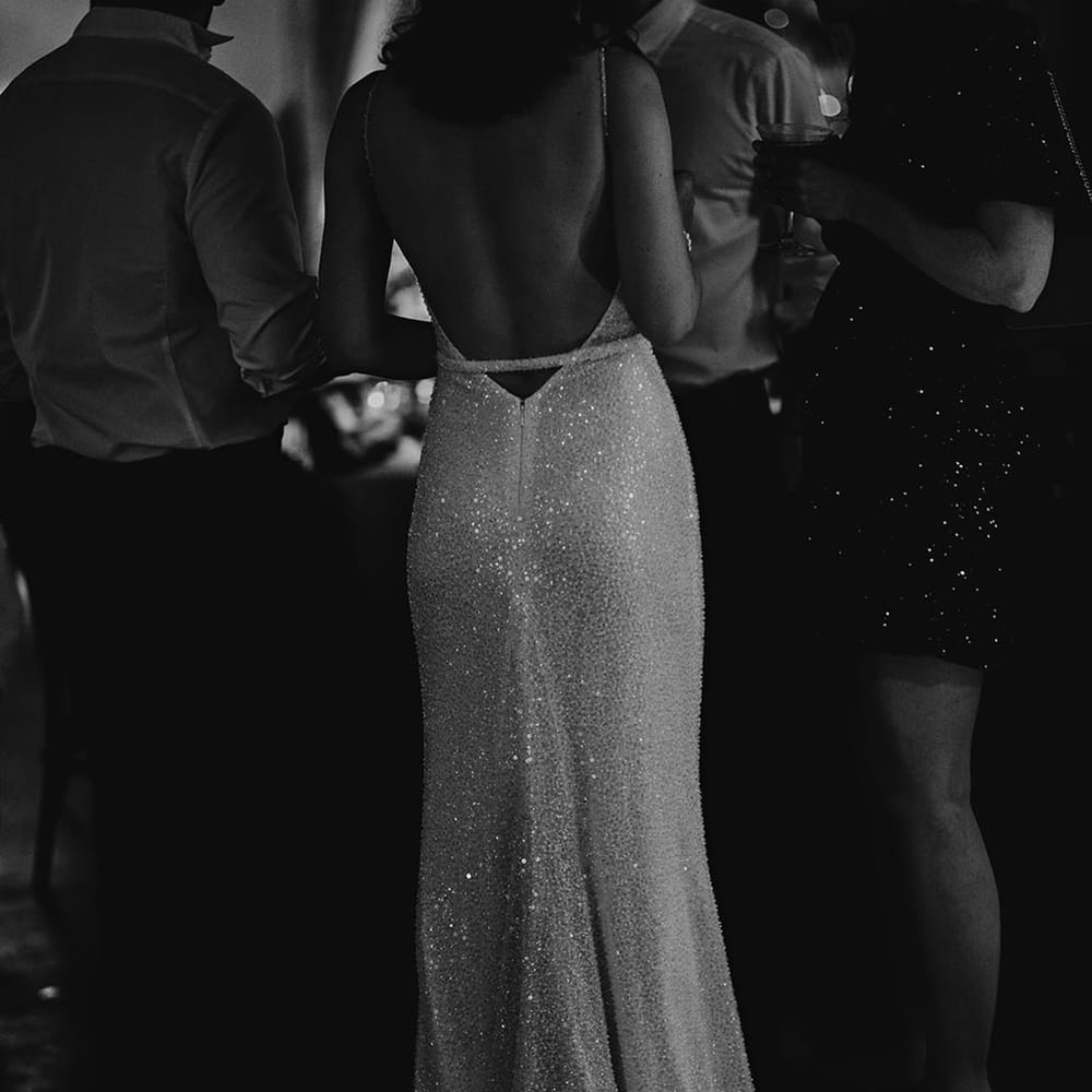 The bride stands socialising in a backless beaded wedding dress from Made With Love Bridal 