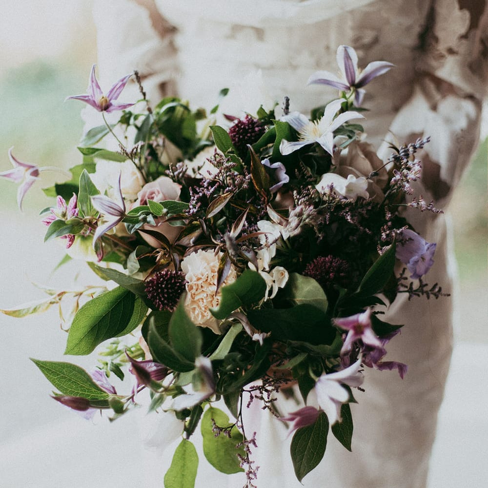 Bride in long sleeve lace detail dress holding mixed floral bouquet with white carnations, purple tulips, roses and foliage