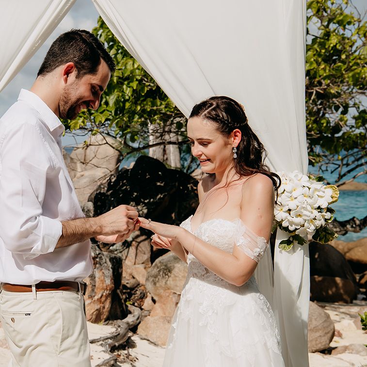 Groom in crisp white shirt, light coloured chinos and tan leather belt putting wedding ring on bride wearing off the shoulder lace wedding dress with sweetheart neckline