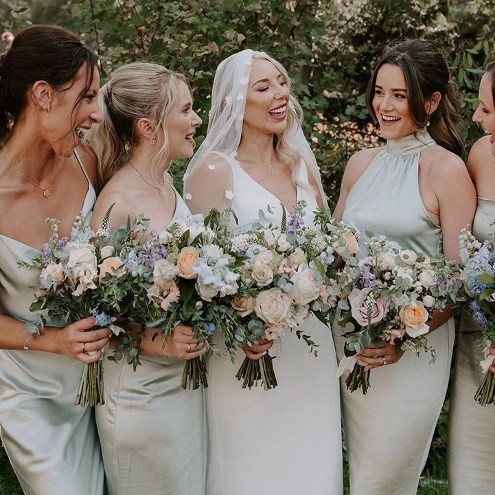 Bride in flower applique veil laughs with the bridesmaids in sage green dresses holding pastel bouquets 
