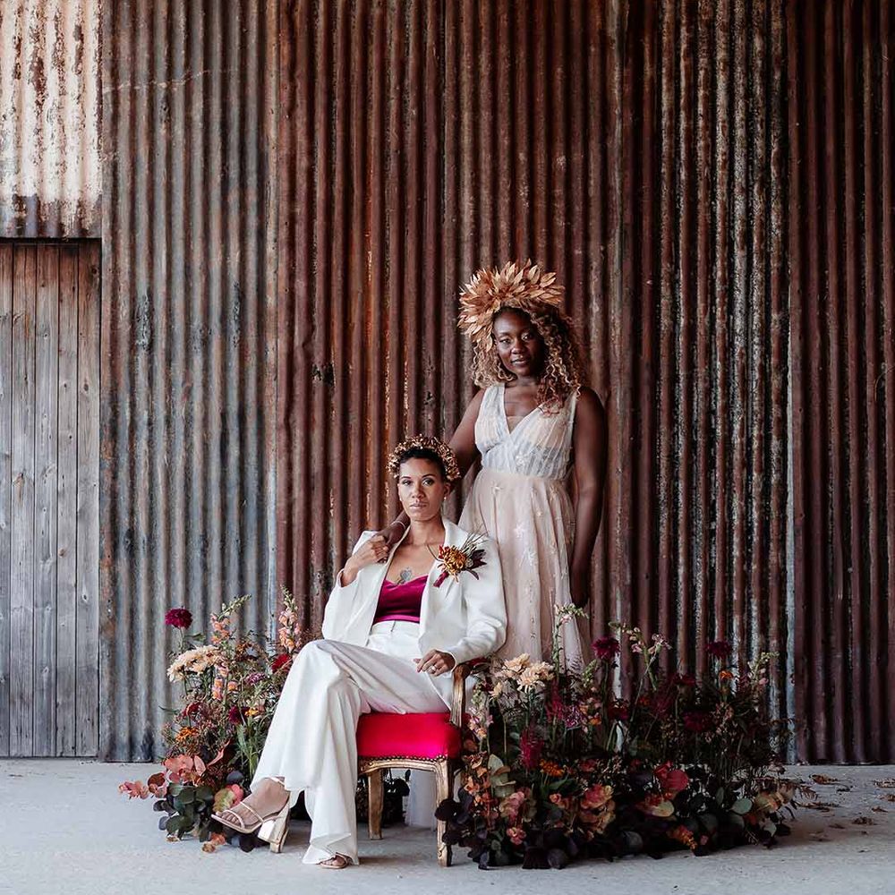 Bride in sleeveless floaty peach gown with embroidered stairs and gold leaf crown standing with bride sitting on velvet and gold chair wearing white silk suit with autumnal dried floral boutonniere and gold bridal crown 