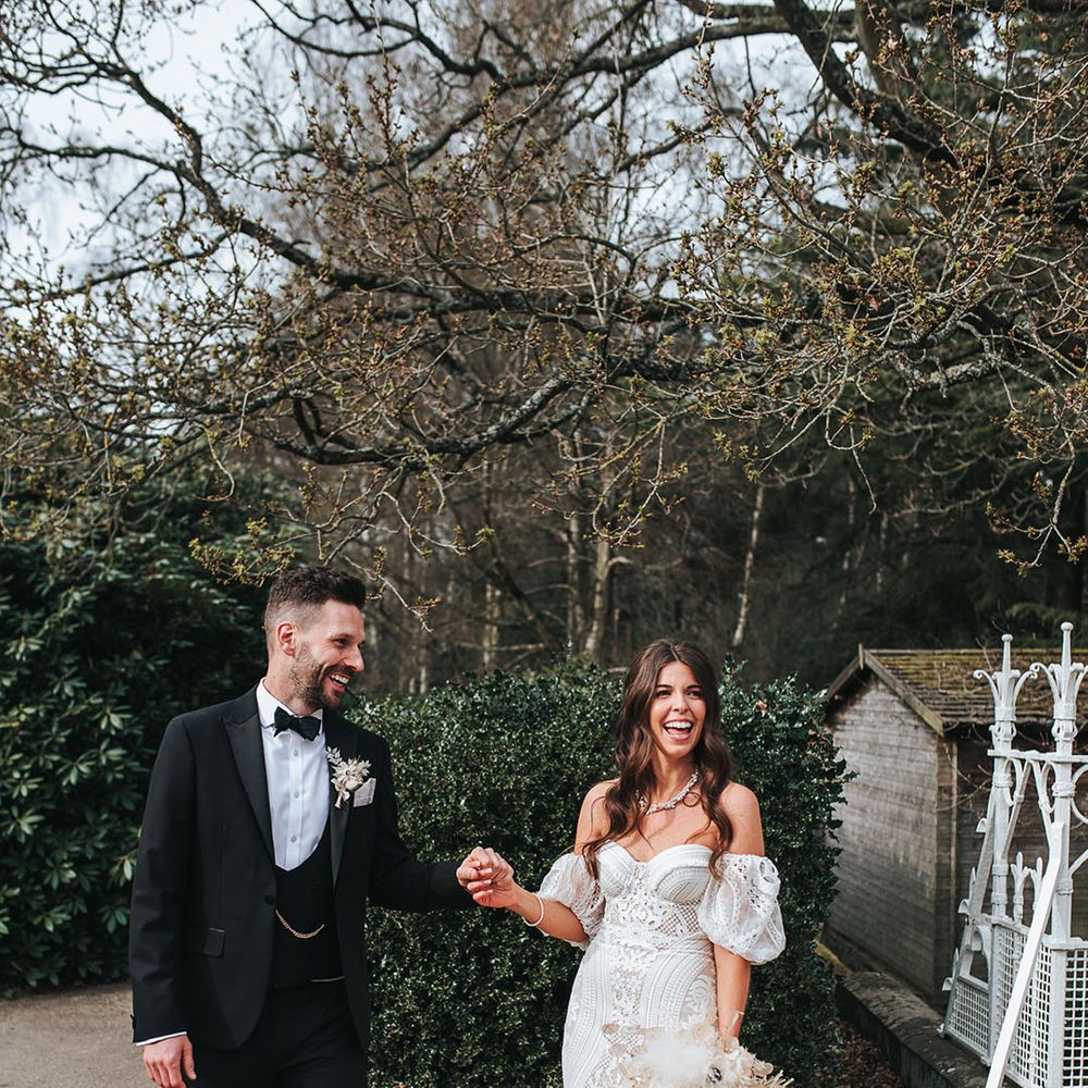 Groom in three piece black tux holding hands with the bride in a boho fitted lace dress for their wedding 