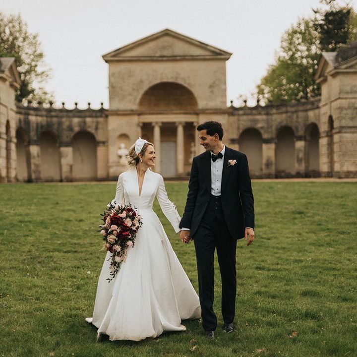 The groom in black tie smiles at the bride as they walk holding hands together with the bride carrying her peony and rose cascading bouquet