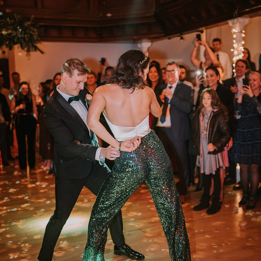 Bride in low back detail taffeta crop top, dark green sequin trousers, chunky velvet green platform heels and bridal crown dancing with groom in classic black tux 