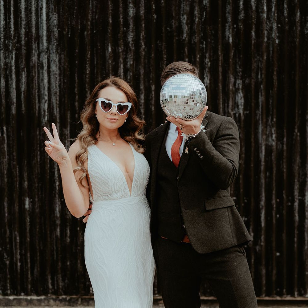 Bride in sequin patterned Made With Love wedding dress with white retro wedding sunglasses with the groom in dark suit holding disco ball decor over his face 