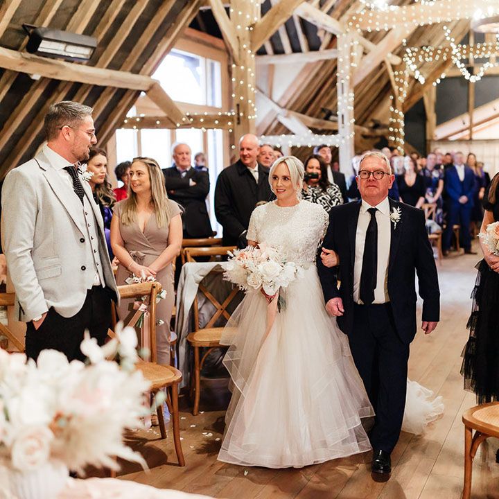 The father of the bride in black and white suit walks the bride down the aisle to the groom for the rustic barn wedding 