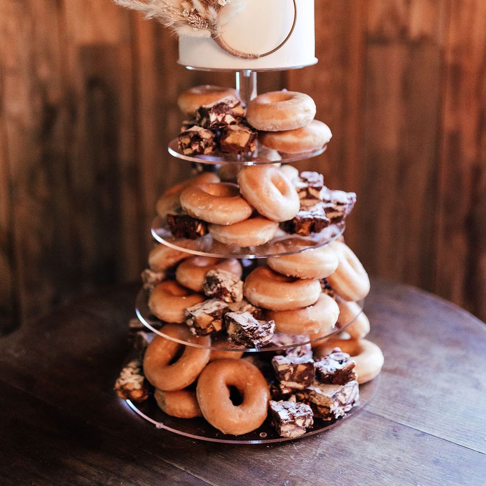 Glazed and iced doughnuts on acrylic cake stand with layer of white frosted cake at the top of the stand with pampas grass decoration