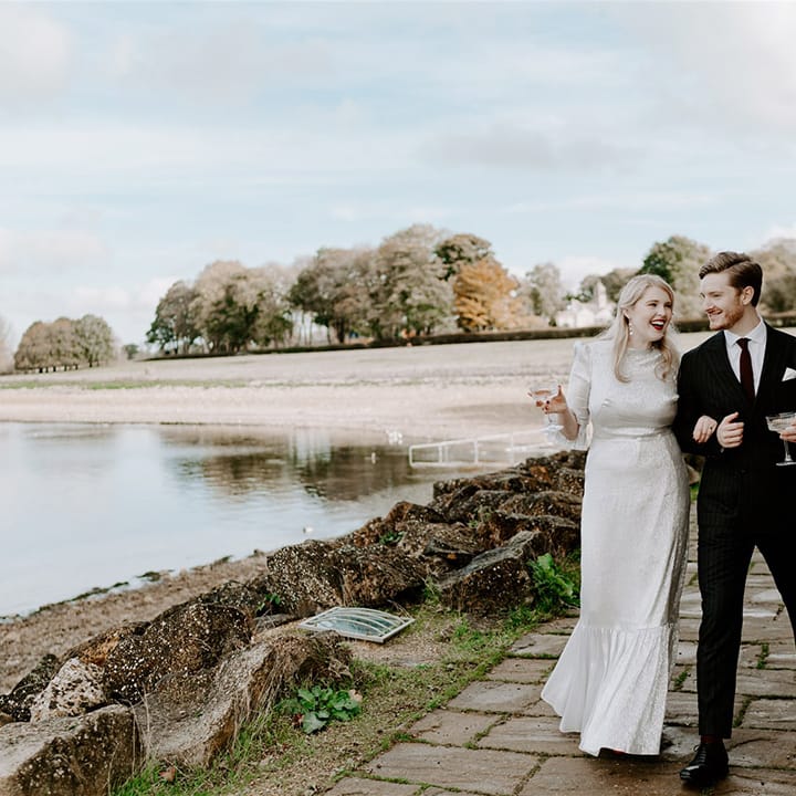 Bride in silver wedding dress walking with the groom in a pinstriped suit drinking champagne from coupe glasses 