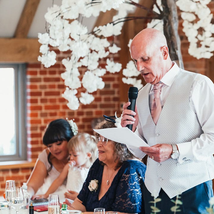 Father of the bride in a light grey waistcoat and pink tie reading out his speech at the reception 