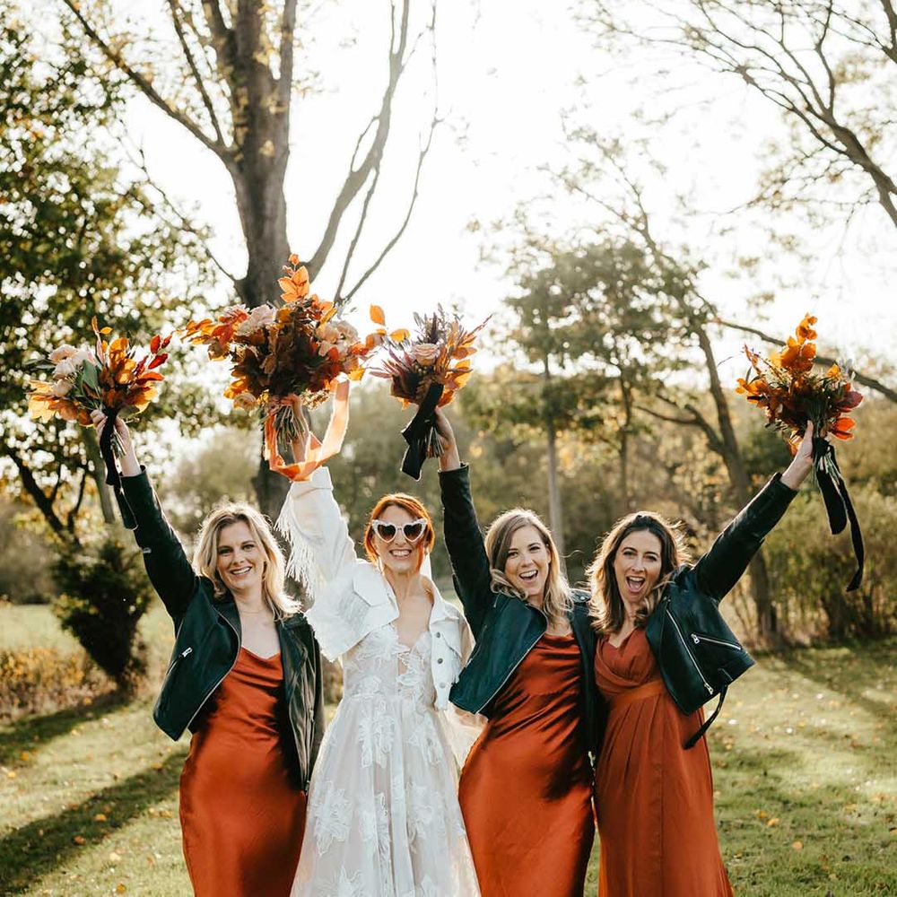 Bride in 3d applique leaf wedding dress with white denim jacket bridal coverup standing with bridal party in burnt orange bridesmaid dresses and black cropped jackets 