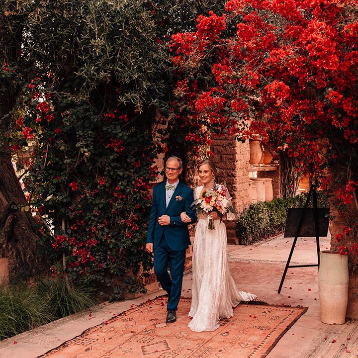 Father of the bride in blue suit and colourful tie walking the bride in modern lace bridal two piece down the aisle to the wedding ceremony 