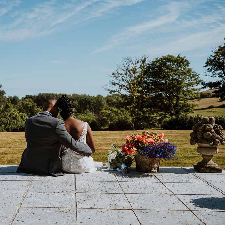 Bride in v back lace and mesh wedding dress sitting with groom in charcoal grooms suit on the steps of Tredudwell Manor wedding venue looking out at the grounds and gardens 