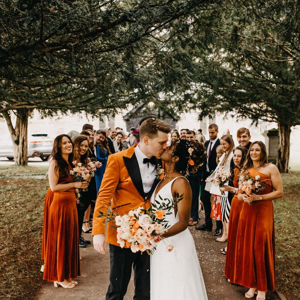 Groom in crushed burnt orange velvet grooms blazer, black bowtie and black suit trousers kissing bride in v neck sleeveless wedding dress with 3D floral applique train with bridesmaids in one shoulder burnt orange bridesmaid dresses and groomsmen in classic black grooms suits at Brinsop Court wedding