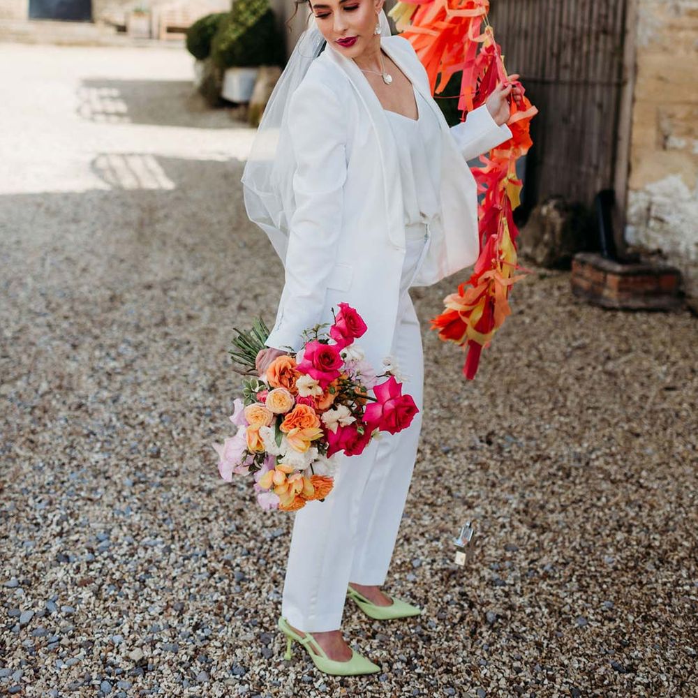 Bride in white bridal suit with closed toe bright green wedding heels holding large balloon decoration with wedding streamers attached outside of Merriscourt wedding venue