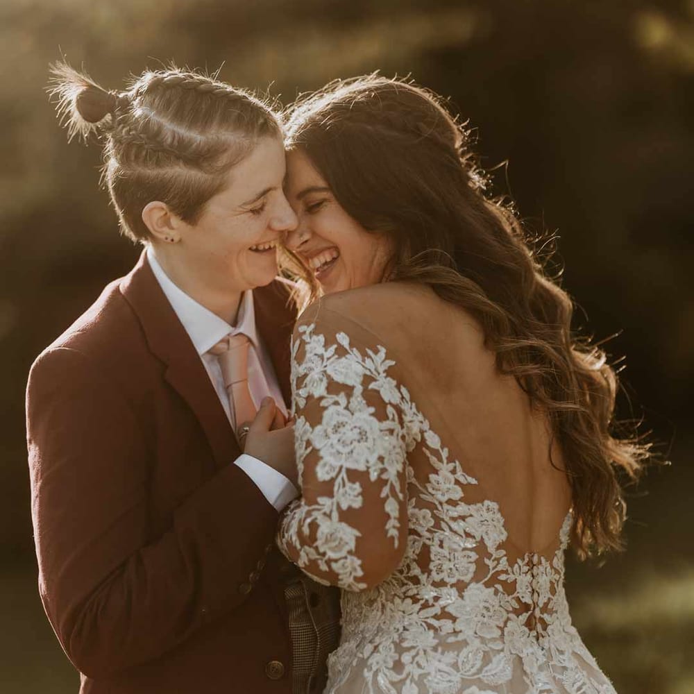 Bride in pretty open back illusion sleeve wedding dress at golden hour embracing bride in burgundy suit 