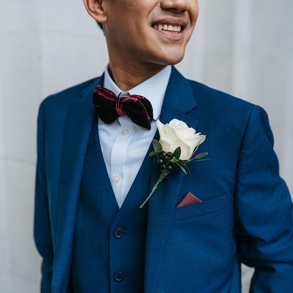 Groom smiles and looks back wearing a blue suit with maroon bow tie and floral buttonhole