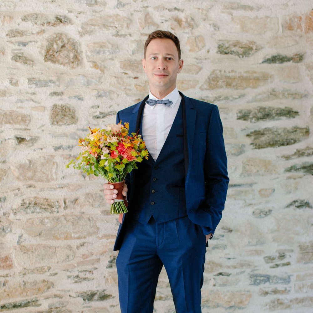The groom holding a bouquet, wearing a dark blue three piece suit