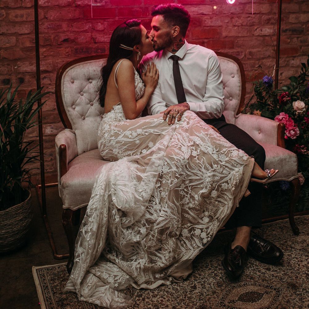 Bride in a lace wedding dress cosying up to her husband on an ornate sofa under a personalised pink neon wedding sign 