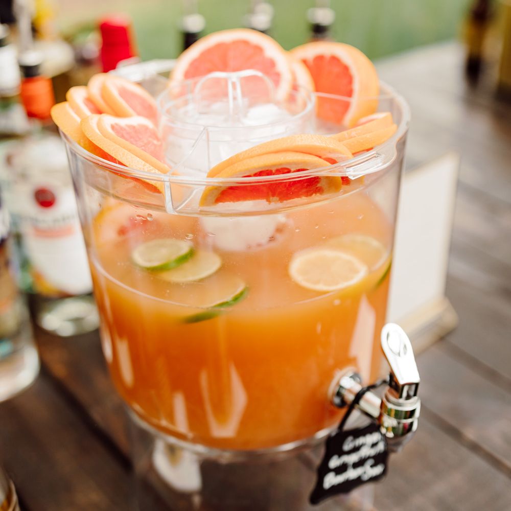 Drinks dispenser full of orange liquid and various citrus fruits on wooden table at backyard wedding