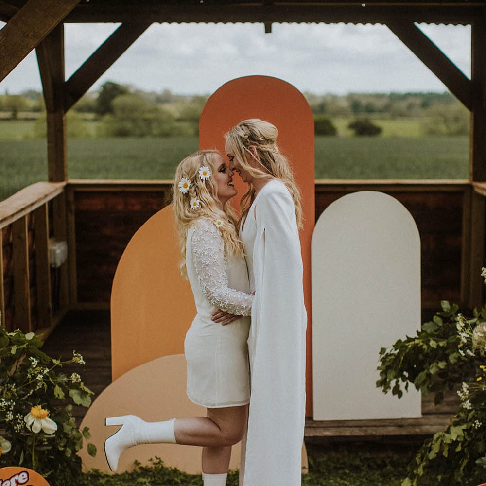 Bride in a mini wedding dress with applique long sleeves and white boots kissing her bride in a wedding jumpsuit at an outdoor sixties wedding dress