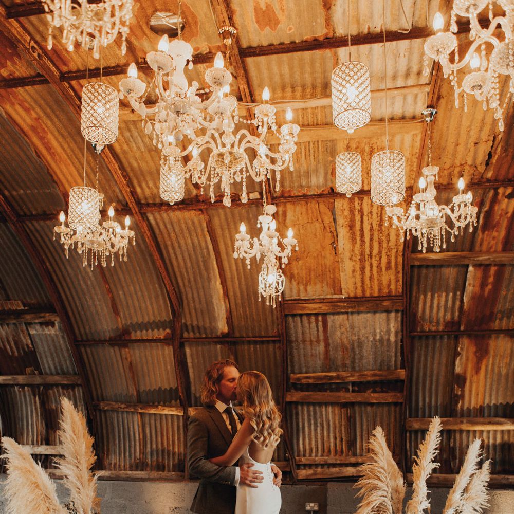 Bride in white Made With Love wedding dress with open back, button detailing and train kisses groom in sage green suit under lit chandeliers surrounded by woven baskets filled with pampas grass and dried leaves in the Hut at Anran Devon