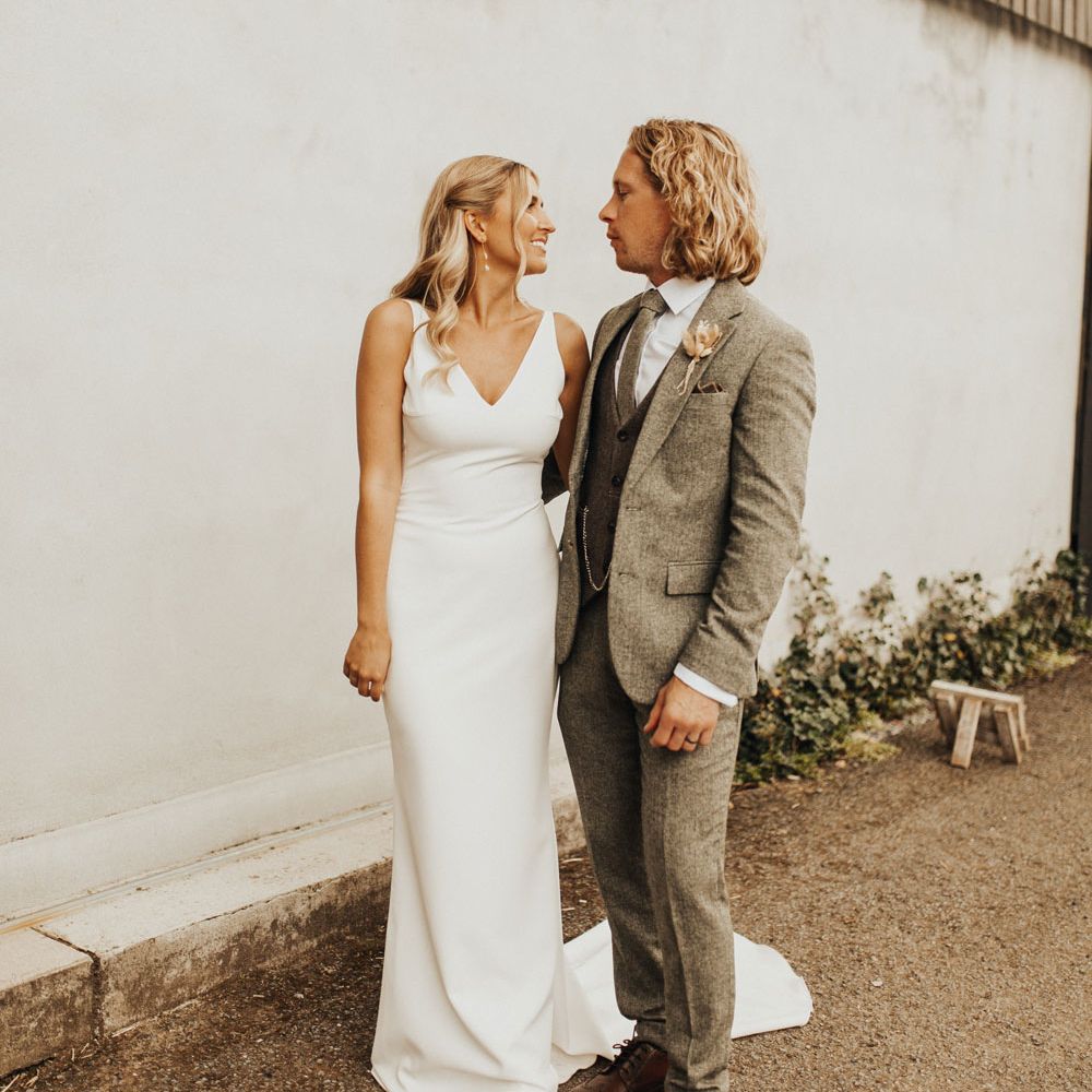 Bride in white v neck Made With Love wedding dress and groom in sage green three piece suit and brown shoes stand smiling at each other outside at Anran Devon