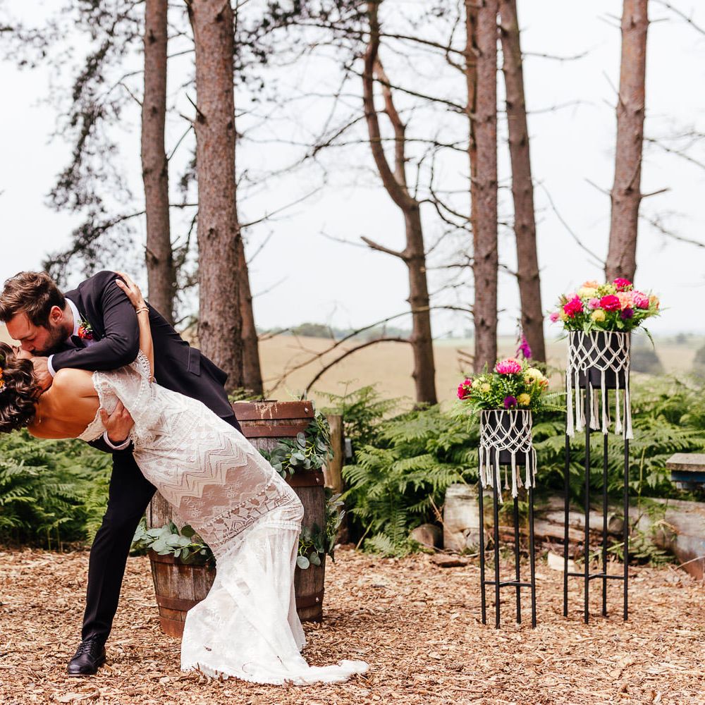 Bride in Grace Loves Lace wedding dress and flower crown is held by groom in black suit for first kiss during woodland wedding ceremony