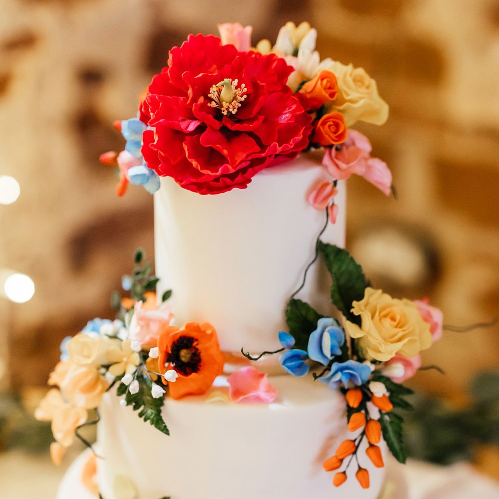 White three tier iced wedding cake with multicoloured floral decorations on wooden table at The Bridal Barn wedding reception