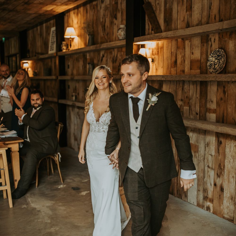 Groom in three piece brown suit with white rose buttonhole walks through barn wedding reception holding hands with bride in lace wedding dress