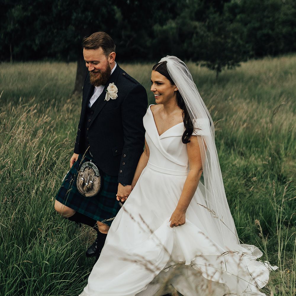 Bride in a Suzanne Neville Bardo wedding dress and long veil walking through the fields with her groom in a tartan kilt 