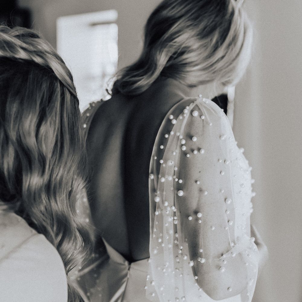 Bride in pearl wedding dress with sleeves has her dress buttoned up by bridesmaid before wedding
