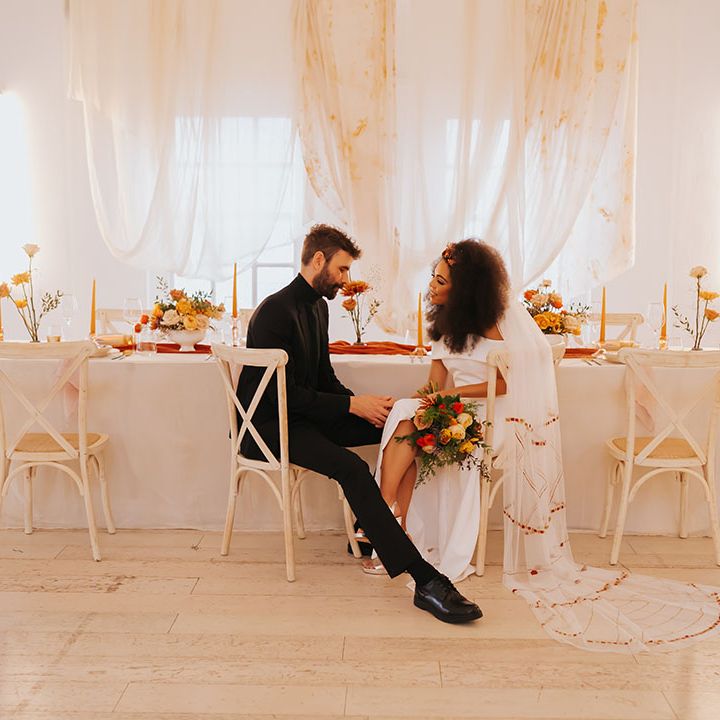 Black bride in an off the shoulder wedding dress with patterned veil sitting at her reception table with her groom in a all black suit and top 