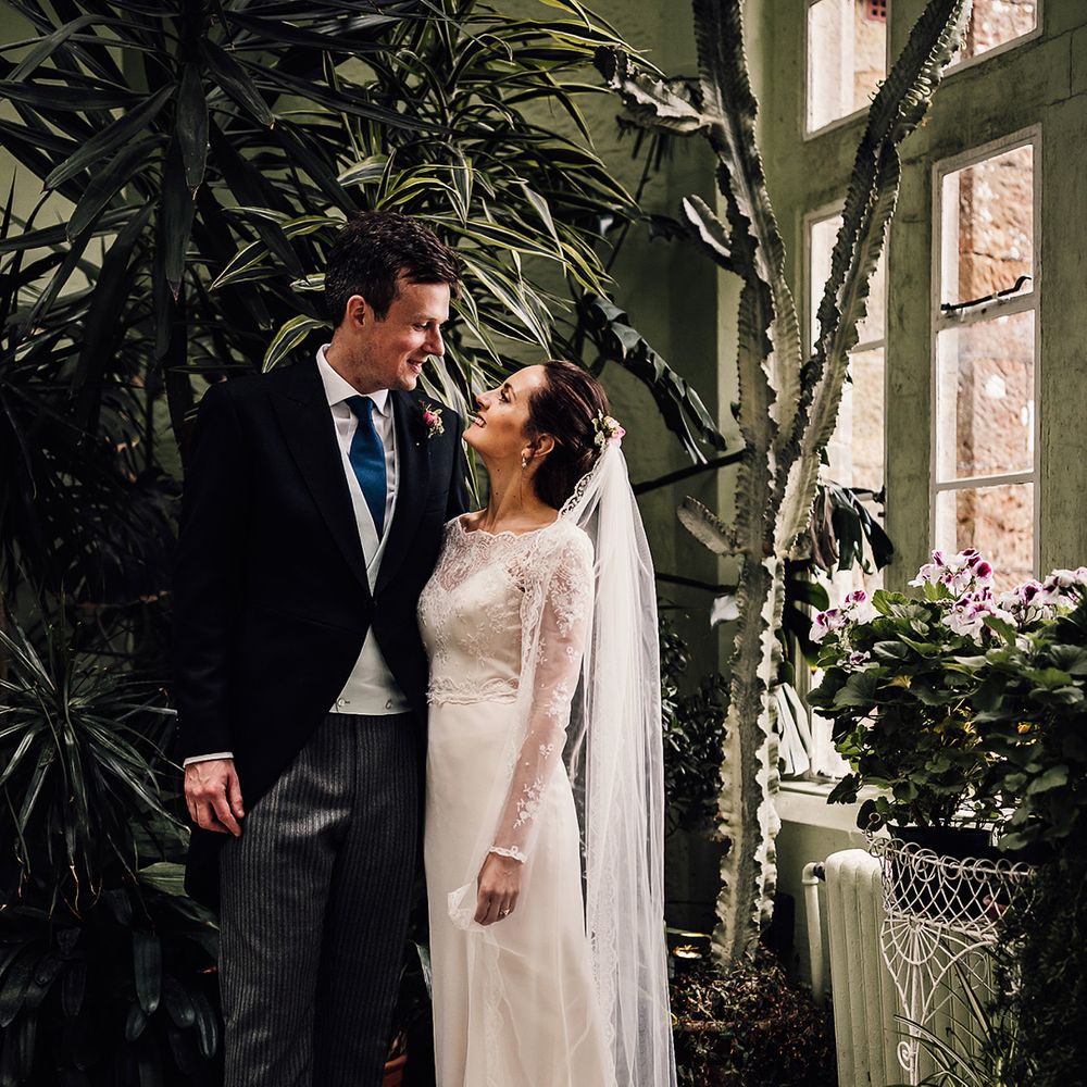 Groom in morning suit with blue tie and grey waistcoat holding bride in satin wedding dresses with sheer lace sleeves and lace embroidered veil 