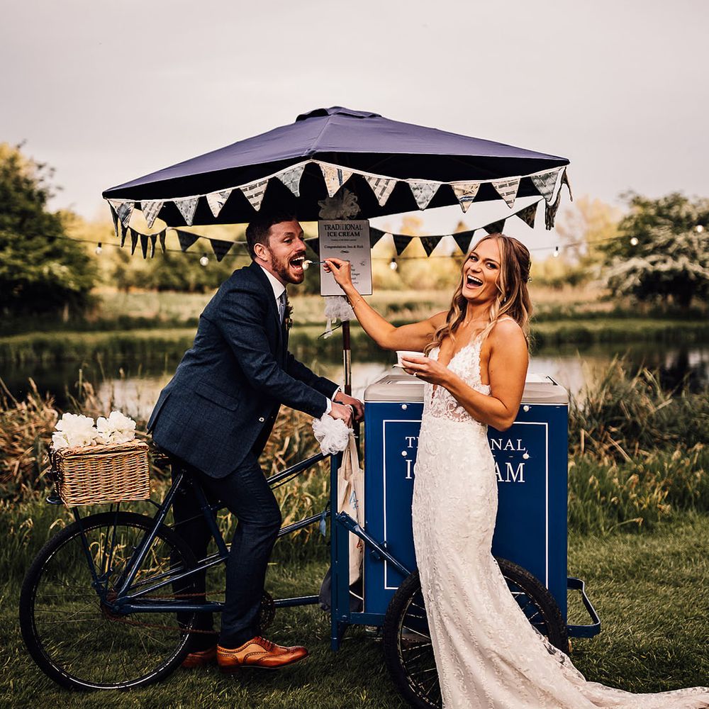 Groom sits on the bike as the bride stands and feeds him ice cream 