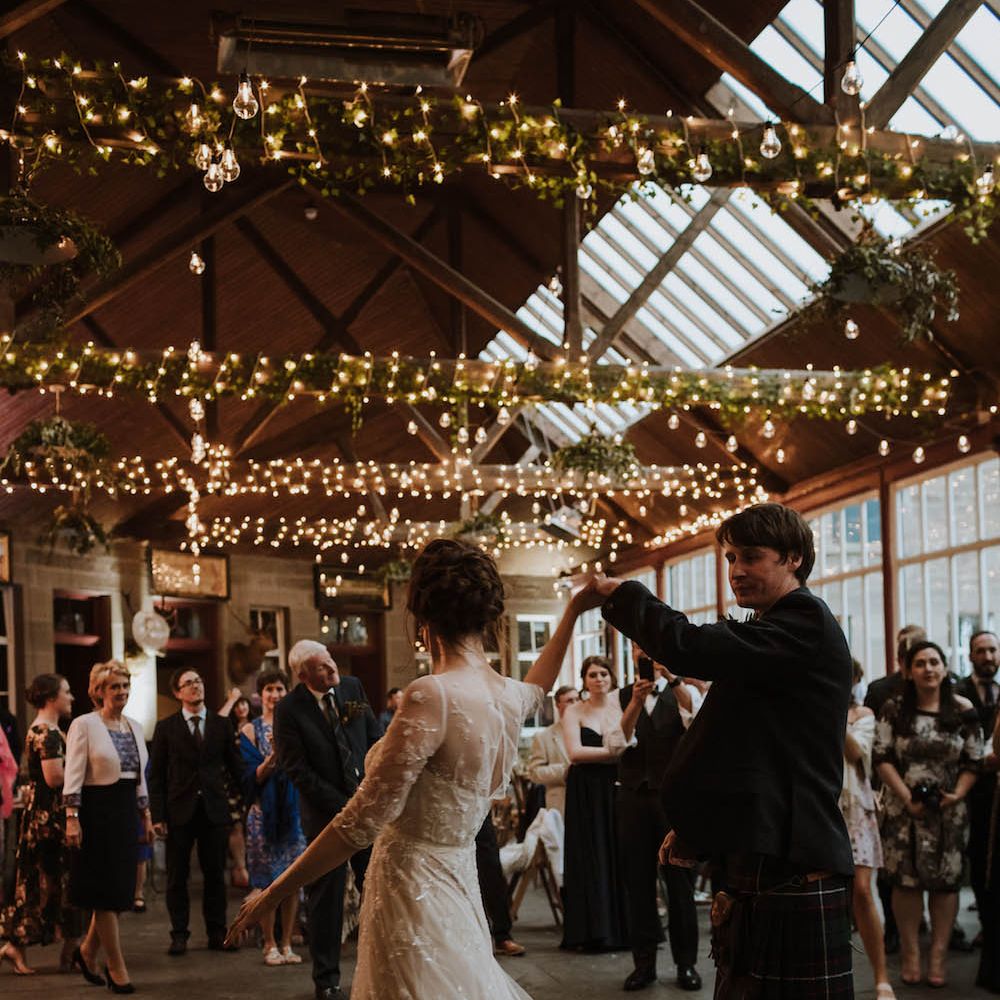 Groom in a kilt twirling his bride in a lace dress at Errol Park wedding venue with fairy lights wrapped around the beams 