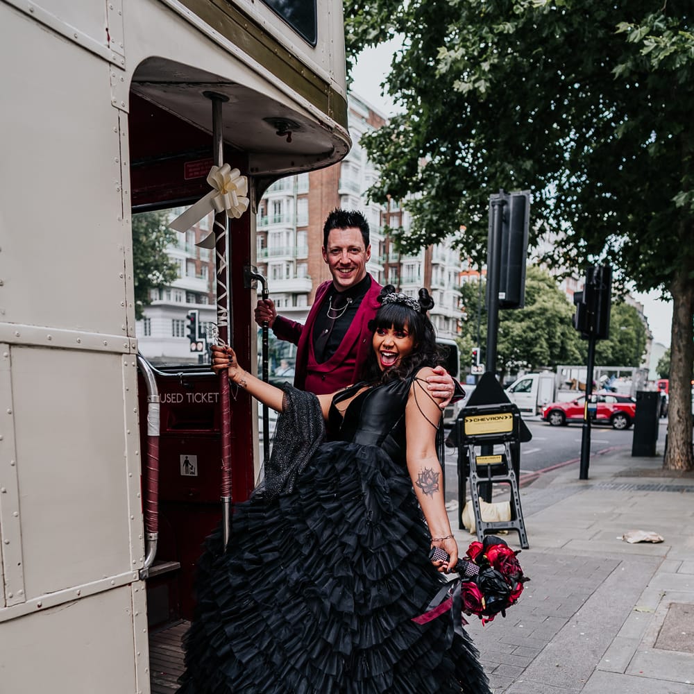 Bride holds red and black rose bridal bouquet and stands with her groom beside white retro London bus 