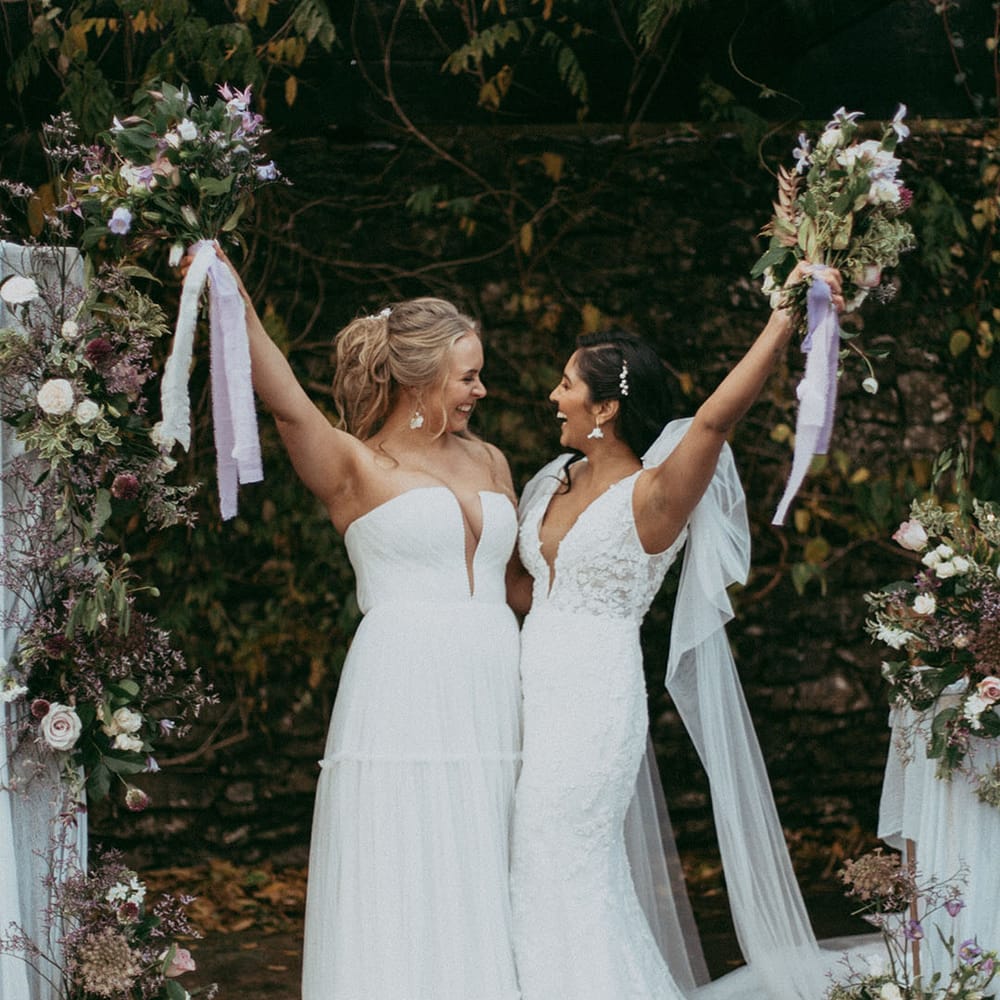 Brides in short sleeve v neck wedding dress with puff lace shoulders and sleeveless ruched wedding dress holding up mixed floral bouquets with with white carnations, purple tulips, roses and foliage and lilac and white lace ribbon