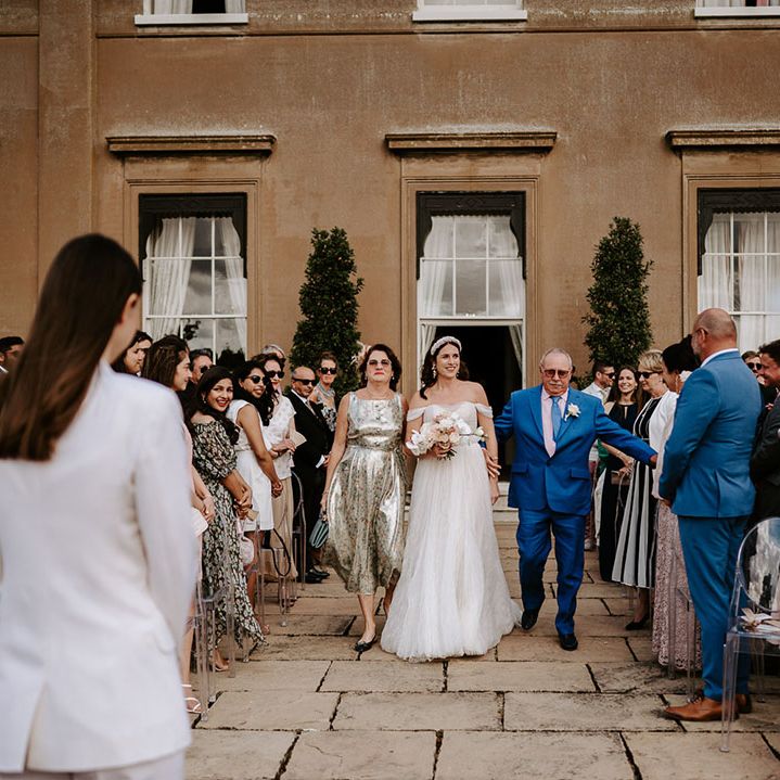 Bride in off the shoulder wedding dress is walked down the aisle by her mother and father with the bride waiting at the end of the aisle 