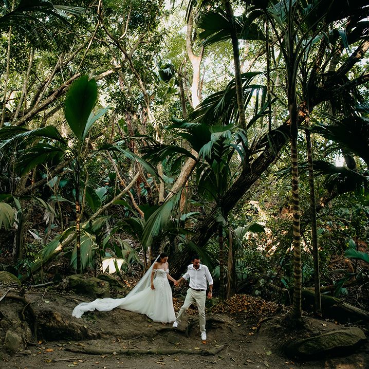 Bride in off the shoulder wedding dress with lace floral details and cathedral length veil holding hands with groom in white shirt, light chinos and white trainers walking through forest in Seychelles
